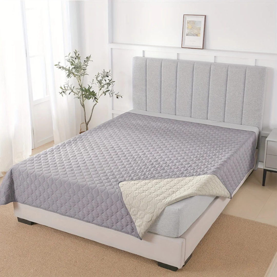 Gray quilted bedspread on a bed in a bedroom setting with a plant and framed picture in the background.